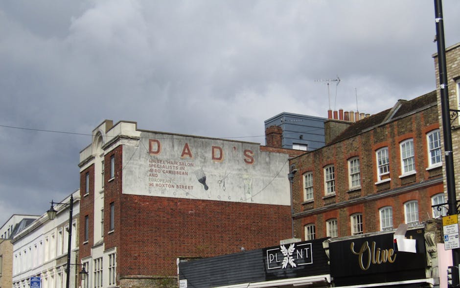 A view of a multi-story brick building with a faded white sign reading 'DAD'S' painted on the upper section. The sign features weathered red lettering and additional, less legible text below. The building's exterior combines red bricks with some areas of white paint or plaster. Adjacent structures include other brick buildings, with a part of a black awning with white text visible at street level, along with a small black sign displaying the word 'OLIVE.' The scene is set against a cloudy, overcast sky, which casts diffuse natural light over the urban environment. The surrounding area appears to be a city street with commercial properties, and the overall atmosphere conveys a typical urban setting that could be related to private waste handling or local rubbish removal, fitting within the context of maintaining cleanliness and addressing fly-tipping outside residential or commercial premises.