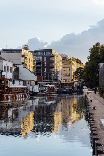 A canal scene in Paddington featuring modern multi-storey residential buildings in the background with a variety of textures and finishes, including glass balconies and dark facades. The canal water reflects these buildings and the partly cloudy sky above, creating a mirror-like surface. On the right side of the image, a paved walkway runs alongside a low curb, with potted yellow bollards placed intermittently along the edge. To the left, several boats are moored, with one boat having a wooden exterior and visible ropes. The scene appears to be captured during late afternoon or early evening, with soft natural lighting highlighting the reflective water and the urban environment. The lush green foliage of trees adds a natural element to the scene, emphasizing the contrast between city living and nature, and subtly illustrating an area where local rubbish collection or private waste disposal might be relevant. This setting aligns with the context of independent waste handling solutions such as those offered by Rubbish Clearance Paddington, especially in urban waterways and residential areas requiring on-site clearance or alternative rubbish removal methods.