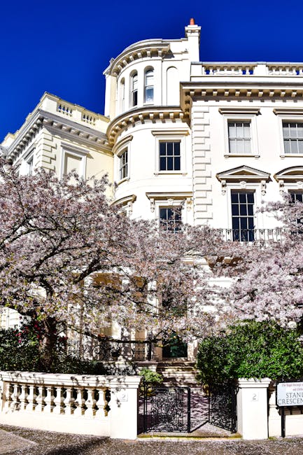 A close-up view of a grand, white Victorian-style building featuring ornate architectural details, including curved bay windows and decorative cornices, with the facade partially obscured by blooming cherry blossom trees in soft pink and white flowers. The building is situated on a paved street, with a small gated entrance visible in the foreground, and lush green bushes bordering the property. The bright blue sky in the background highlights the building's clean, freshly painted exterior, and the overall scene suggests a well-maintained residential property. In the context of rubbish removal services, this setting may imply the importance of efficient, private waste disposal for high-value, historic properties, and the scene subtly aligns with the concept of professional rubbish clearance in well-preserved urban environments, such as Paddington, where premium estate clearance services might be required to maintain aesthetic standards.