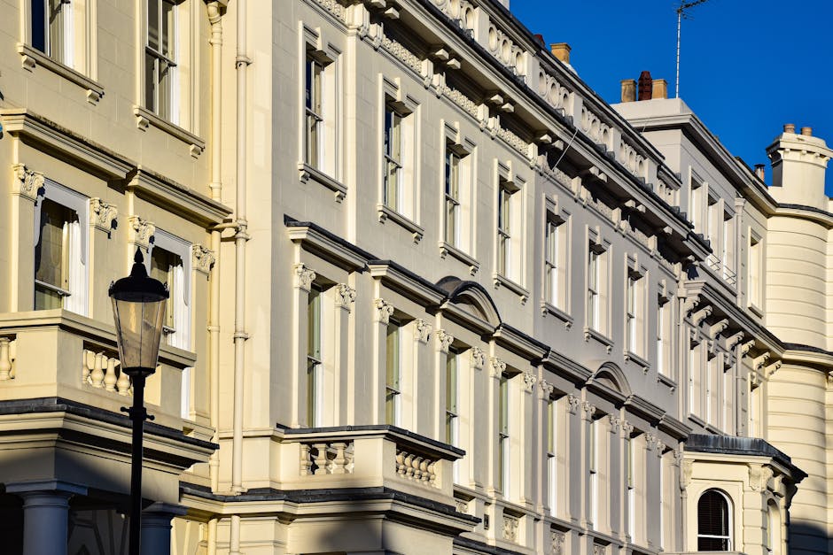The image shows a close-up view of a historic multi-storey building with ornate architectural details, including decorative mouldings, cornices, and window surrounds, painted in a cream or off-white colour. Several large windows are visible, some with open sash panes, framed by detailed trim and small ledges. Vertical and horizontal lines from the building’s structure create a grid pattern across the façade, highlighting its classical design. In the foreground, a traditional black street lamp is positioned, indicating an urban setting, with a bright blue sky serving as the backdrop, suggesting a clear, sunny day. The overall scene reflects a well-maintained, historic street environment, commonly associated with areas where private waste collection or rubbish clearance services such as Rubbish Clearance Paddington operate to manage waste disposal outside residential or commercial properties, particularly in maintaining the aesthetic integrity of such ornate buildings. The detailed exterior and urban context subtly relate to the theme of managing rubbish for refurbishment or estate clearance purposes.