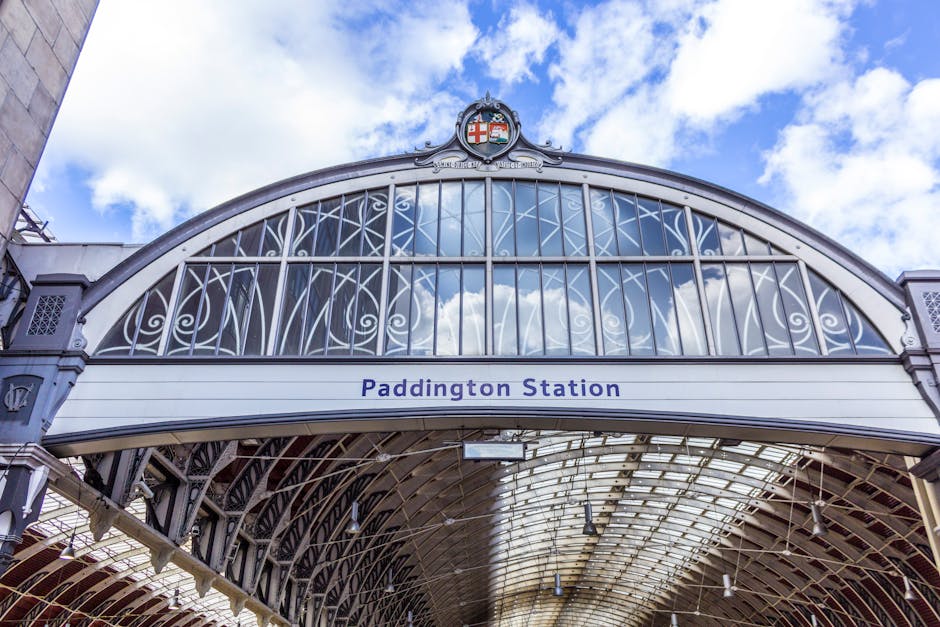 The image shows the exterior view of Paddington Station's ornate metal archway, which features intricate white decorative ironwork in a lattice pattern, supporting a large curved glass canopy. The arch is topped with a crest displaying the Union Jack and other heraldic symbols. Below the arch, a white panel bears the station name 'Paddington Station' in dark blue lettering. The glass canopy allows natural daylight to illuminate the station entrance, with the sky visible in the background, showing a mix of white clouds and blue sky. The surrounding structures include parts of the station's facade, with visible steel framework and decorative architectural details. This setting highlights the historical design elements typical of railway station architecture and provides a context for urban transportation hubs. The image captures a clear, well-lit scene that emphasizes the station's grand entrance and its characteristic Victorian-era design, relevant to discussions on urban infrastructure and heritage preservation within the scope of waste management and infrastructure maintenance services.