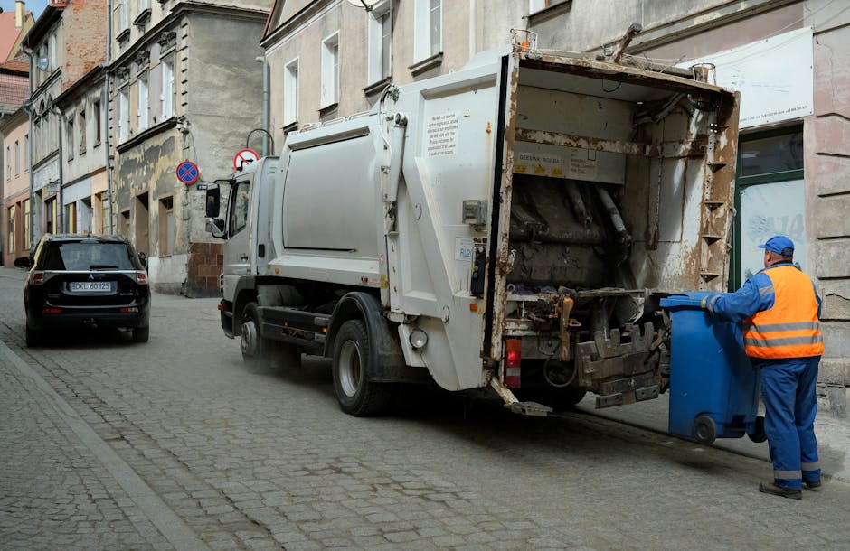 A waste collection truck operated by Rubbish Clearance Paddington is parked on a narrow urban street with cobblestone pavement, positioned next to a block of older, weathered buildings with graffiti marks and a mix of plaster and brick facades. The truck's rear compartment, made of metal with visible signs of rust and wear, is open, revealing evidence of recent rubbish collection activities. A worker dressed in high-visibility orange and blue safety clothing, including a helmet and gloves, is loading a blue plastic bin into the truck's hopper, which features mechanical components adapted for compacting waste. In the background, a black car is parked on the opposite side of the street, and prominent street signs indicating parking restrictions are mounted on the building wall. The scene is cast in natural light, emphasizing the utilitarian nature of the environment and the practical aspect of private waste disposal services, which often operate independently from traditional local authority rubbish collection approaches.