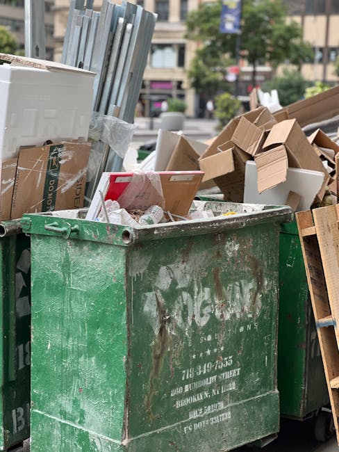 A large, weathered green waste skip filled with various types of rubbish, including crumpled cardboard boxes, broken pieces of white foam packaging, and scattered paper debris, positioned outdoors on a street or public area. In the background, there are multiple cardboard boxes, some flattened and others still in folded form, alongside additional wooden pallets or furniture parts, suggesting recent clearance or disposal activity. The environment appears urban, with blurry trees and buildings visible in the distance, indicating a city street scene. The scene is illuminated by natural daylight, highlighting the textures and colors of the waste materials, which range from the rough, corrugated texture of cardboard to the smooth, glossy surfaces of foam packaging. The presence of the Rubbish Clearance Paddington company’s waste bin in the scene implies an independent collection or on-site clearance as part of a private waste handling service. The image emphasizes the extent of accumulated rubbish ready for disposal, fitting within the context of rubbish removal services and alternative waste management options in the area.