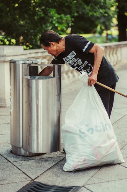 A man with dark hair, wearing a black T-shirt with white text and black trousers, is bending forward on a paved sidewalk while disposing of waste into a clear, metal recycling bin. He is holding a large white plastic bag filled with rubbish in his left hand and using a wooden stick or broom with his right hand to insert waste into the bin. The recycling bin has a cylindrical shape with multiple compartments and a metallic finish, positioned near a low stone or concrete balustrade that runs along a street or park area. In the background, there are lush green trees and foliage, suggesting an outdoor public space with natural light illuminating the scene. The pavement tiles are arranged in a regular pattern, and the overall environment appears clean and well-maintained. This image illustrates a discreet, outdoor waste disposal activity aligned with rubbish removal services typical of independent collection or environmental management efforts by local groups or waste handling providers like Rubbish Clearance Paddington, emphasizing responsible waste disposal in urban settings.