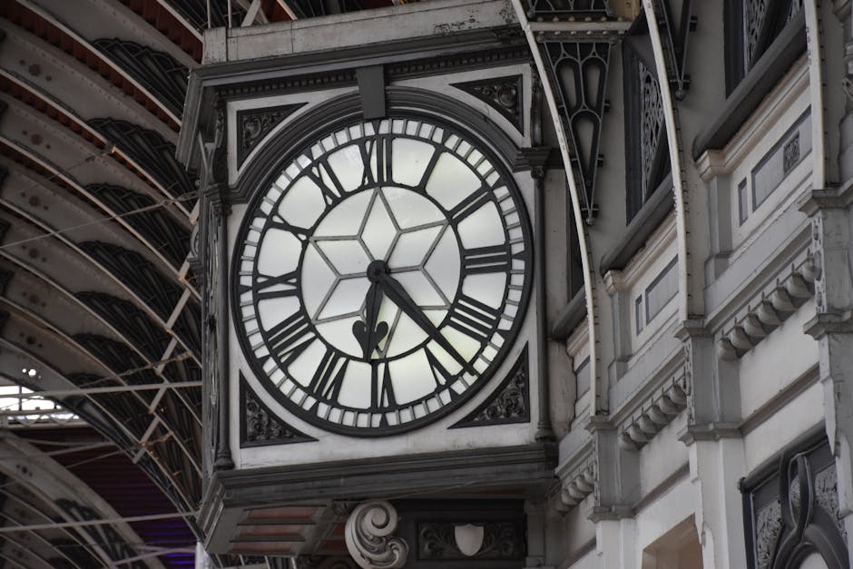 A large, ornate clock with a white face and black Roman numerals is mounted on the exterior of a historic building. The clock features a black minute hand pointing to 7 and an hour hand pointing slightly past 5, indicating the time as approximately 5:35. The clock is enclosed in a decorative black and white frame with intricate carved detailing, including scrollwork and floral motifs, contributing to its classical appearance. To the left and right of the clock, portions of the building’s facade are visible, showcasing detailed white stonework with evenly spaced rectangular panels and decorative cornices. Above the clock, an arched section of the building’s architecture is visible, with curved metal beams and structural elements that appear to support a covered platform or railway station roof. The lighting in the scene suggests daylight, with a soft, natural illumination highlighting the textures and details of the clock and the surrounding architecture, creating a neutral, professional visual context that subtly aligns with themes of public or private historical site preservation without promotional language.