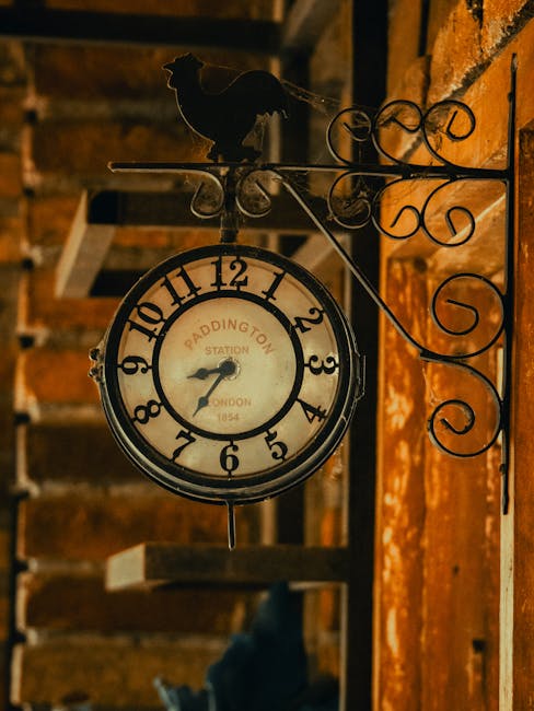 A vintage-style round clock with a white face and black numerals, mounted on a decorative wrought iron bracket affixed to a red brick wall. The clock features black hour and minute hands, with the time showing approximately 7:45. The clock face bears the inscription 'PADDINGTON STATION LONDON 1854'. On top of the clock, there is a black metal silhouette of a dog, positioned facing to the right. The surrounding environment appears to be an indoor or sheltered area with a wooden structure visible in the background, including a wooden staircase or shelving unit. The lighting is warm, casting soft shadows on the brick wall, and the scene conveys an old-fashioned, nostalgic atmosphere that subtly relates to traditional or independent methods of timekeeping, paralleling themes of alternative waste handling and on-site clearance, as managed by companies like Rubbish Clearance Paddington.