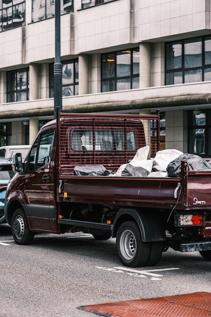 A small, dark red flatbed truck parked on a city street in front of a modern office building with large glass windows and a light-colored stone facade. The truck's cargo area is filled with various rubbish items, including black and white garbage bags, some of which appear to be partially open, revealing waste or discarded materials inside. The truck features protective red metal mesh around the cab area and has a slightly raised tailgate at the rear. The surrounding environment includes a parking space with white painted markings, and a black streetlight pole situated adjacent to the vehicle. The scene is lit by natural daylight, with reflections visible on the building's windows. The image conveys a scene of private waste collection, aligning with a rubbish removal service focused on on-site clearance of various waste materials, such as mattress and household rubbish, through independent collection methods. The context hints at alternative waste handling approaches outside of local authority rubbish collection, consistent with services offered by companies like Rubbish Clearance Paddington.