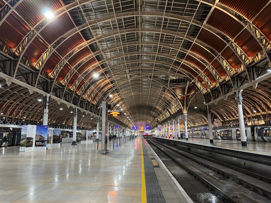 A spacious train station interior featuring a high, arched metal and glass roof with intricate steel framework and warm brown panels. The platform area is illuminated by bright overhead lighting, creating reflections on the polished, light-colored tiled floor. To the right, multiple train tracks are visible, with modern train carriages parked alongside. On the left, display boards and informational signage are mounted on tall metallic poles, with a few passengers seen in the distance. The station appears clean and organized, with a wide empty space in the foreground, subtly indicating the environment where independent waste removal or on-site clearance services like those offered by Rubbish Clearance Paddington could be relevant, especially in maintaining cleanliness amidst bustling travel hubs.