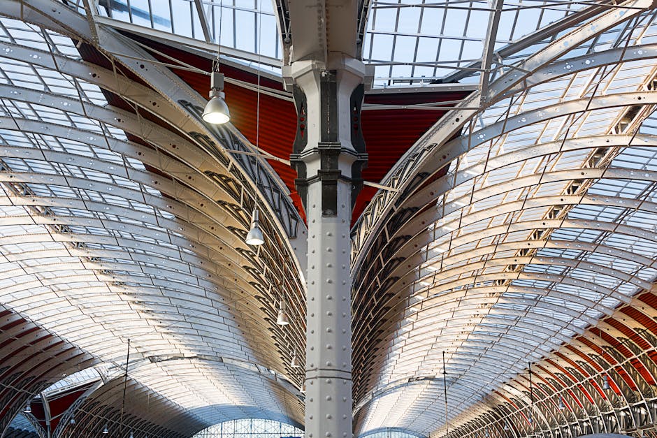 The image shows the interior of a modern architectural structure featuring a large central steel support column with rivets. The ceiling comprises curved, overlapping panels with a metallic finish, some painted in a reddish hue, and is constructed from glass and metal, allowing natural light to filter through. The panels are arranged in a symmetrical pattern that extends outward and upward, creating a vaulted effect. Overhead, several industrial-style pendant lights are suspended from the ceiling, illuminating the area with bright light. The environment appears to be a spacious, possibly commercial or public indoor space with a sleek, contemporary design. In the context of rubbish removal services by Rubbish Clearance Paddington, such a setting might relate to on-site clearance or architectural waste management but in the image itself, only the structural and architectural details are visible without specific objects or debris.