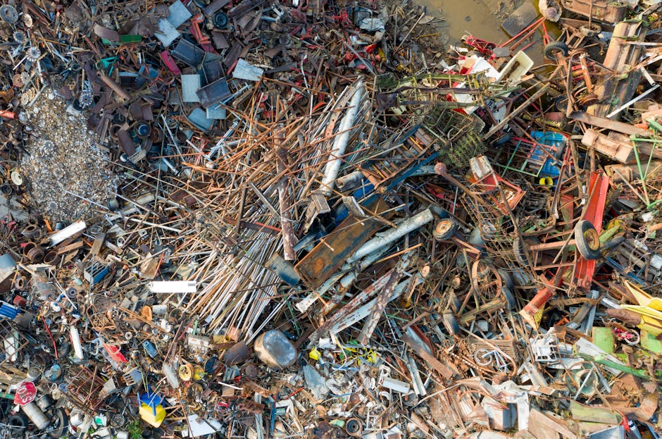 A large, chaotic pile of assorted scrap metal and discarded objects fills the scene, consisting of rusted steel pipes, twisted metal rods, broken bicycle frames, and various damaged appliances. The metal items vary in color from reddish-brown rust and dull silver to faded blue and green paint, with some parts showing corrosion and weathering. The objects are piled on top of each other haphazardly, creating an uneven surface with sharp edges, bent pieces, and tangled wires. In the background, there is a muddy ground with a small amount of standing water, reflecting parts of the debris. Some smaller items, such as plastic containers, bits of wire, and miscellaneous trash, are scattered throughout the heap, adding to the cluttered appearance. This scene likely represents accumulated industrial or household waste awaiting collection or disposal, which could be handled by an independent rubbish removal service like Rubbish Clearance Paddington, highlighting the need for proper waste management solutions.