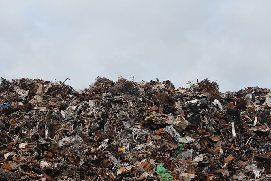 A large pile of mixed waste and debris occupying an outdoor area under a cloudy sky, consisting of various discarded materials such as broken wood, plastic fragments, metal scraps, cardboard, and fabric, all tangled and layered haphazardly. The pile appears to be situated on a paved or compacted surface, with no visible boundaries or containment features. The waste materials show different textures and colors, including brown, gray, black, green, and some bright highlights from plastics or packaging. The overall scene reflects an extensive accumulation of rubbish, typical of outdoor waste disposal sites or private rubbish removal operations, as handled by companies like Rubbish Clearance Paddington, illustrating the scale of waste that can be managed through professional rubbish services or alternative waste handling arrangements. The environment suggests an open area dedicated to debris accumulation, with natural light diffusing over the scene, emphasizing the disorderly mixture of materials and the logistical challenge of waste disposal or clearance.