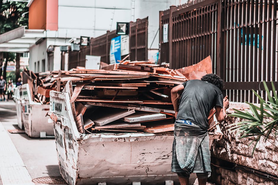 A man with curly black hair, wearing a dark t-shirt and plaid shorts, stands next to a large, white, metal skip filled with rusty, broken roof tiles and construction debris. The tiles are reddish-brown with a rough, uneven surface and are stacked haphazardly, spilling over the sides of the skip. The skip is positioned on a paved sidewalk adjacent to a tall, dark brown metal fence with vertical slats. In the background, there are street signs, a lamppost, and a building with a white exterior and an orange section visible on the side. The environment appears to be an urban area during daylight, with natural lighting and some muted shadows cast across the scene. This image exemplifies an independent waste collection scenario, with private disposal during a renovation or building project, aligning with rubbish removal services provided by companies like Rubbish Clearance Paddington. The overall atmosphere suggests an organized, yet cluttered construction waste disposal situation close to a busy street.